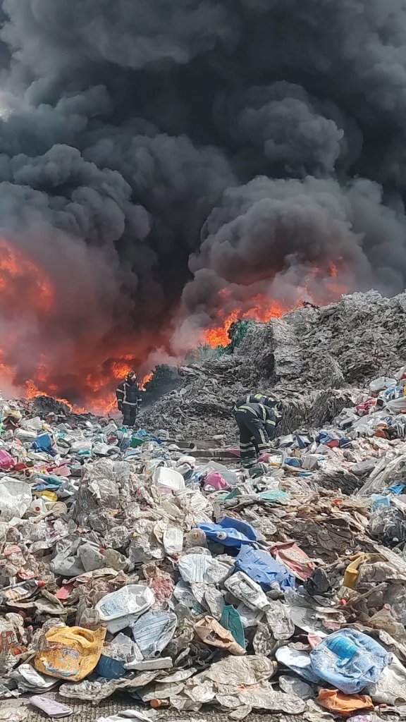 Se puede ver el fuego sobre las pilas de PET mientras los bomberos luchan por apagarlo.